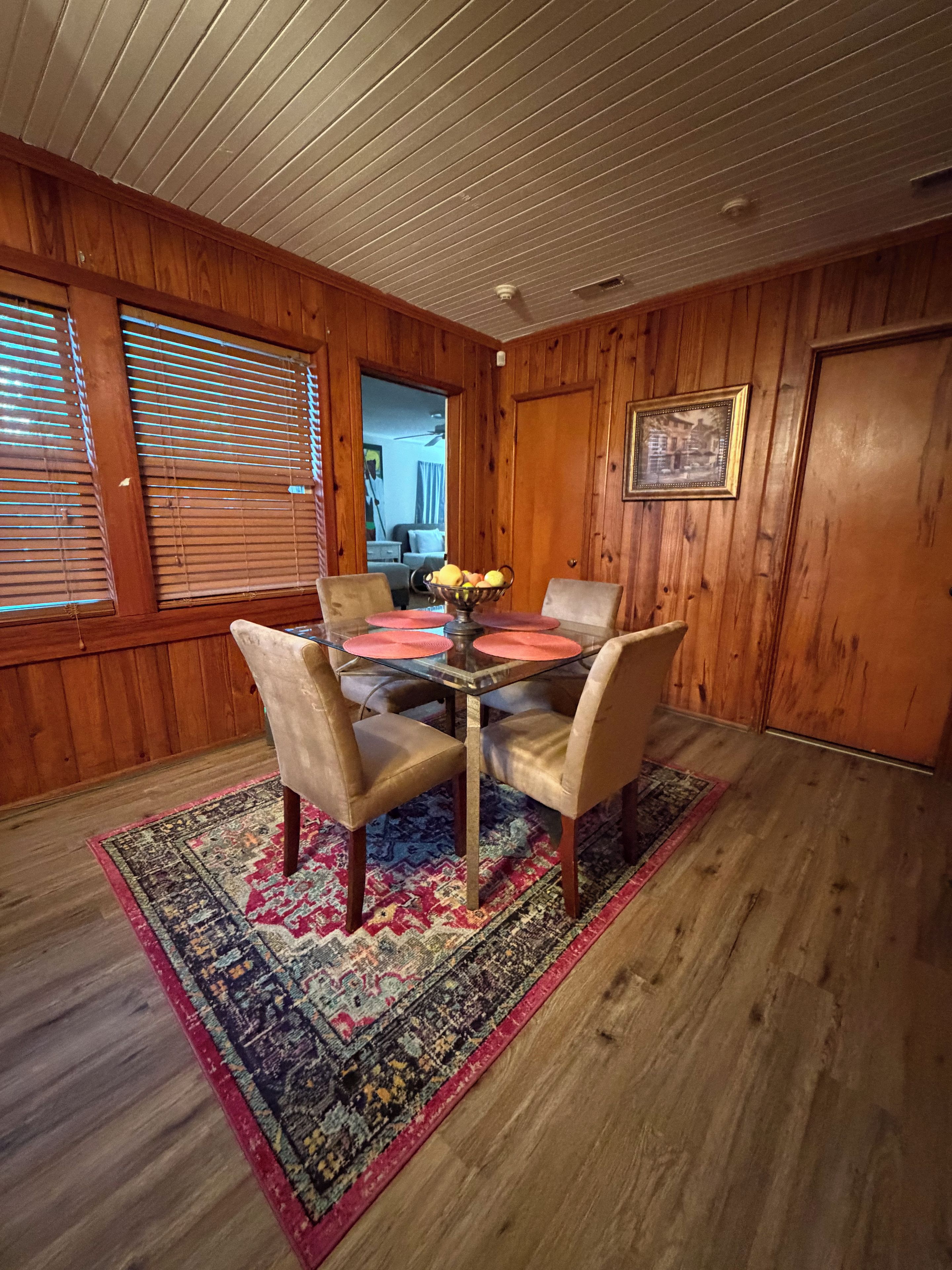 A dining area with a wooden paneled wall, a round glass table with a fruit bowl, four chairs, and a patterned rug.