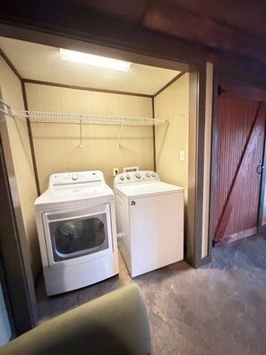 A laundry room with two washing machines and a dryer, a metal shelf above, and a wooden door to the right. - Private 3BR Home • 4 Queen Beds + 3 Sofa Beds • Great Location (Valdosta)