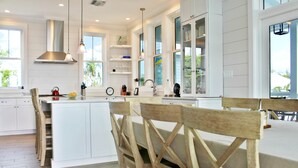 A modern kitchen with white cabinetry, a central island, wooden chairs, and large windows.