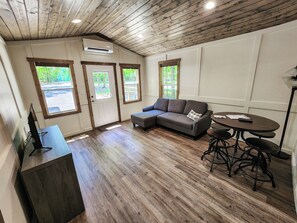 Ein Zimmer mit Holzboden, einem grauen Ecksofa, einem runden Tisch mit Stühlen und einem Fernseher auf einer Holzkommode.