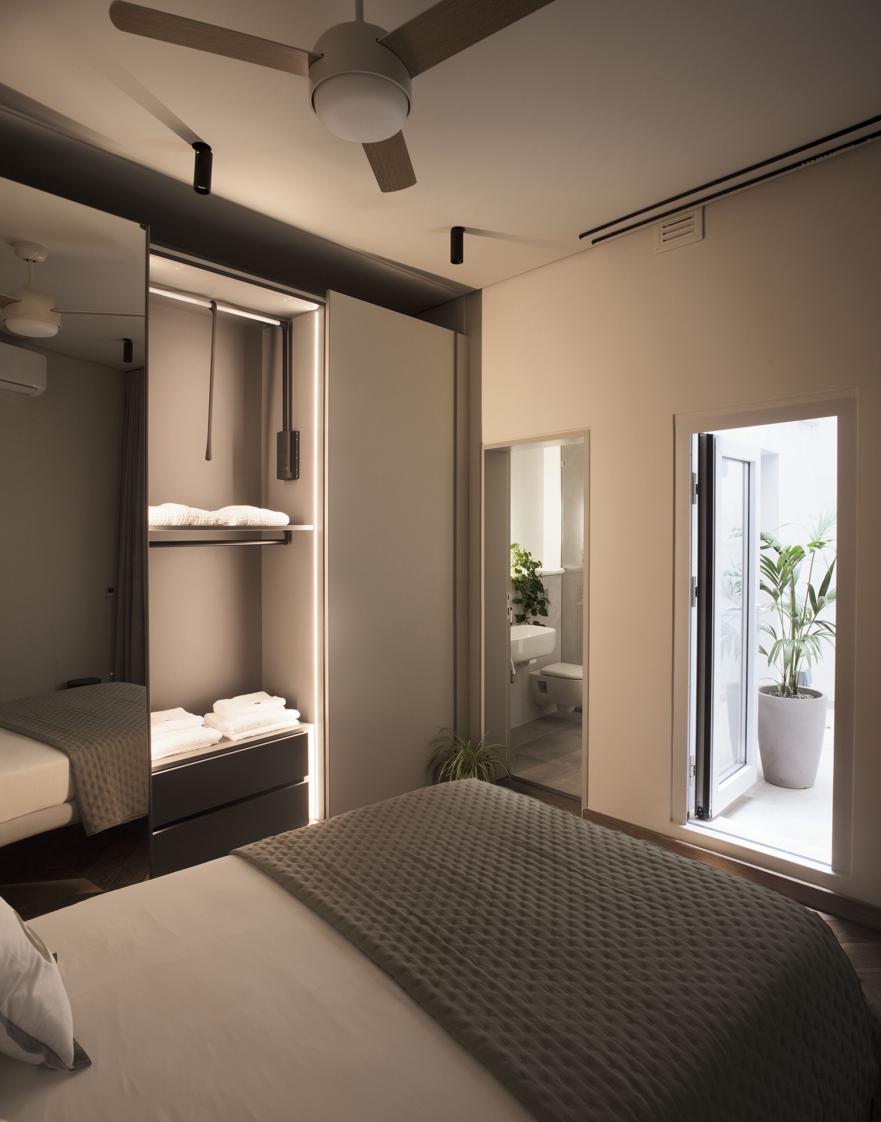 A modern bedroom with a large bed, a wardrobe, a ceiling fan, and a bathroom visible through an open door.