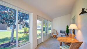 A sunlit patio with a wooden table and chairs, large windows, and a TV. - Spacious 2BR Villa | Pools | Near Beach & Dining | Garage Parking (Port Orange)