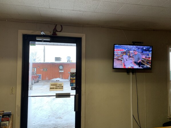 A room with a television mounted on the wall displaying a kitchen scene, a door leading outside, and a sign on the door.
