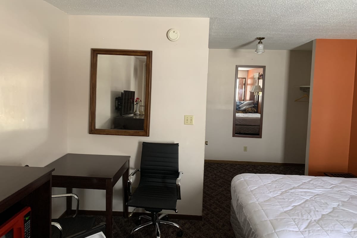 Quarto de hotel com cama, escrivaninha, cadeira, espelho e mesa pequena.