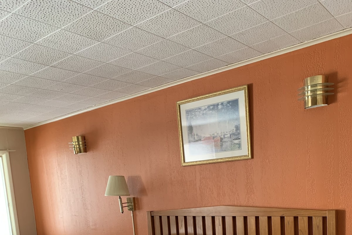 Quarto de hotel com uma cama grande, cabeceira de madeira, quadro na parede, abajur e uma escrivaninha com cadeira.