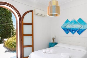 A hotel room with a bed, a nightstand, a wall-mounted air conditioner, and a large window with a view of greenery.
