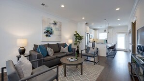 Ein modernes Wohnzimmer mit einer Couch, Sesseln, einem Couchtisch und einem Essbereich mit Stühlen. Im Hintergrund befindet sich eine Küche mit Schränken und einem Kühlschrank.