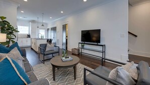 Ein modernes Wohnzimmer mit Sofa, Couchtisch und TV-Möbel. Im Hintergrund befindet sich eine Küche mit Schränken und einem Fenster.