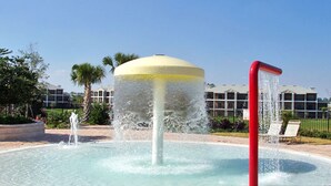 Una fuente de agua con parte superior amarilla y caño rojo, rodeada por una zona pavimentada con una piscina.