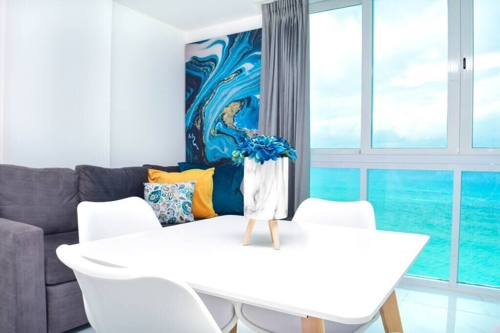 A modern living room with a white table, grey sofa, and a large window with a view of the ocean.