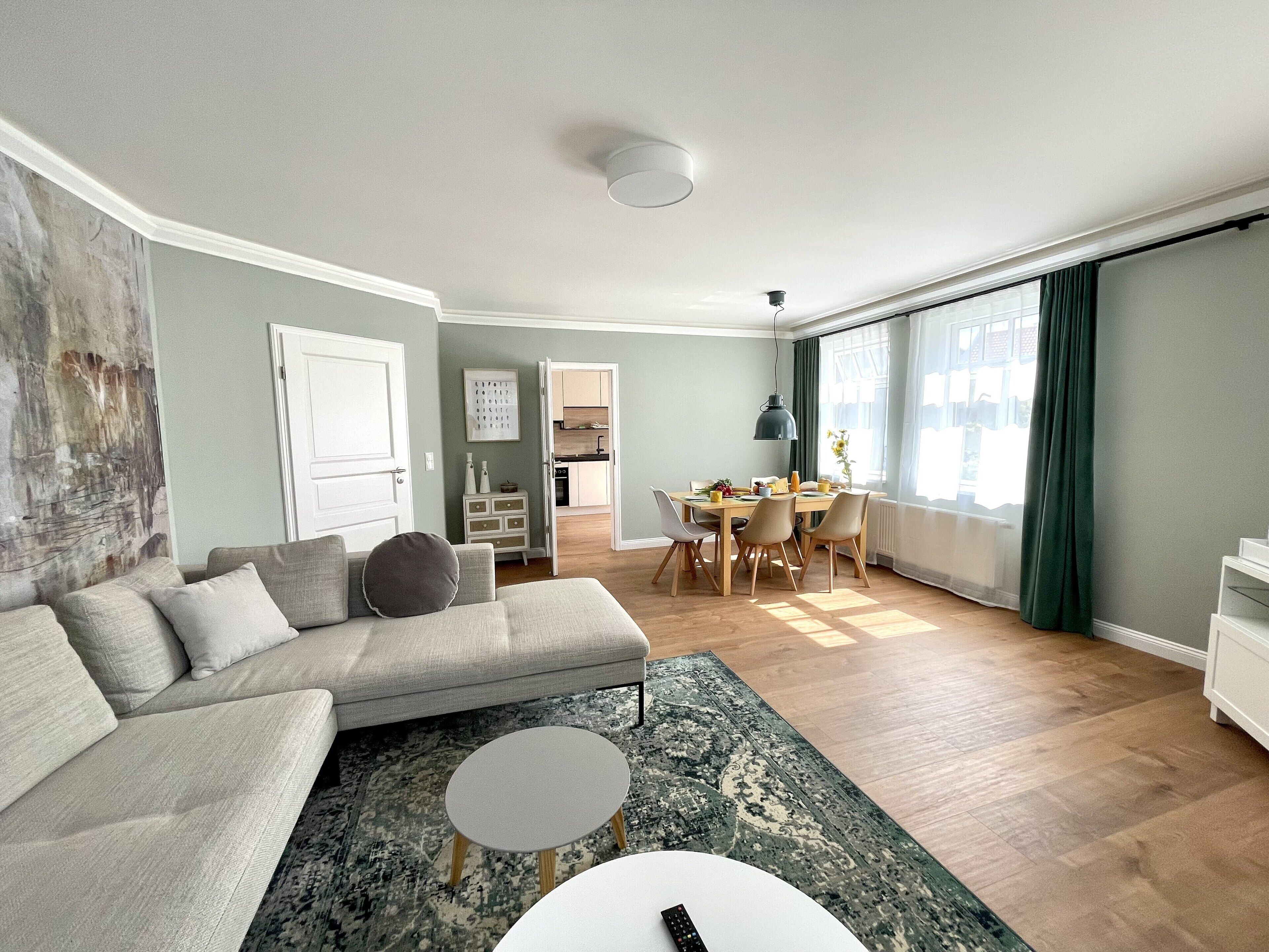 Ein modernes Wohnzimmer mit einer grauen Couch, einem Esstisch und einer Küche im Hintergrund.