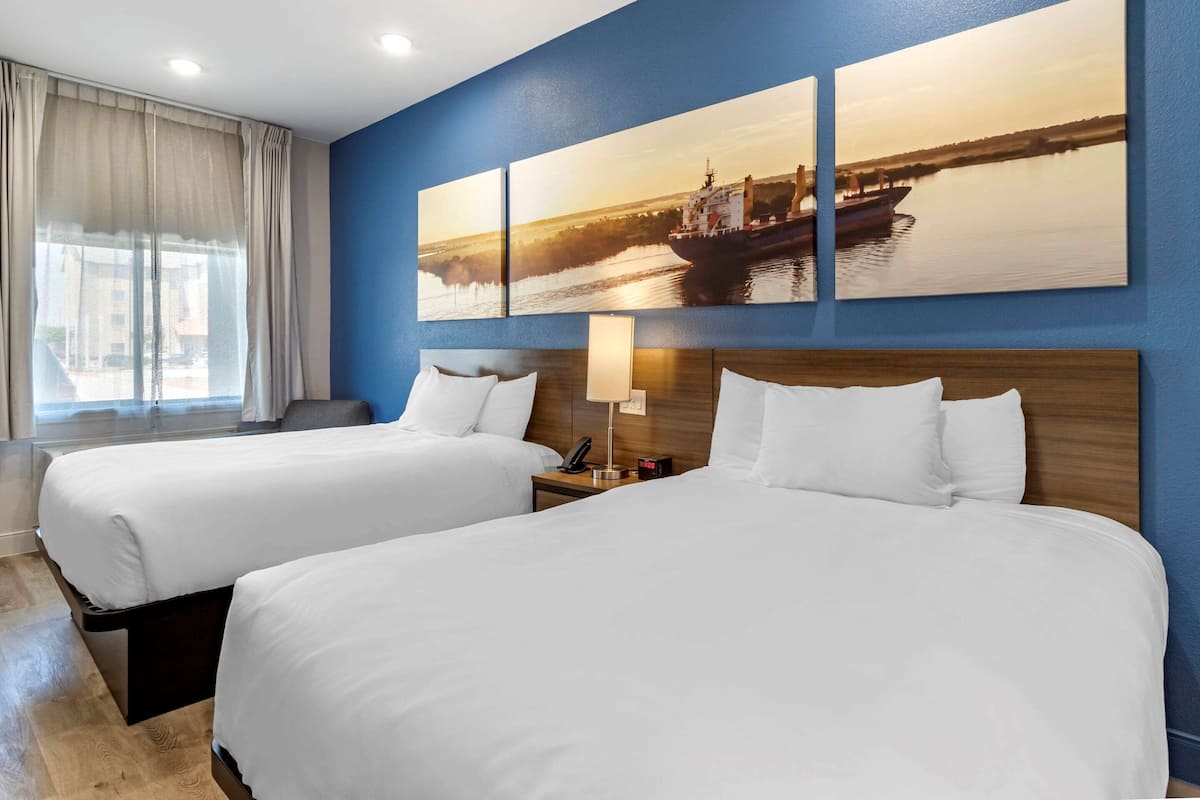 Ein Hotelzimmer mit zwei Betten, einem großen Fenster mit Vorhängen, einem Wandbild mit einer Flusslandschaft und einem Nachttisch mit Lampe und Telefon.
