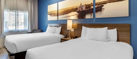 A hotel room with two beds, a large window with curtains, a wall-mounted painting of a river scene, and a bedside table with a lamp and phone.