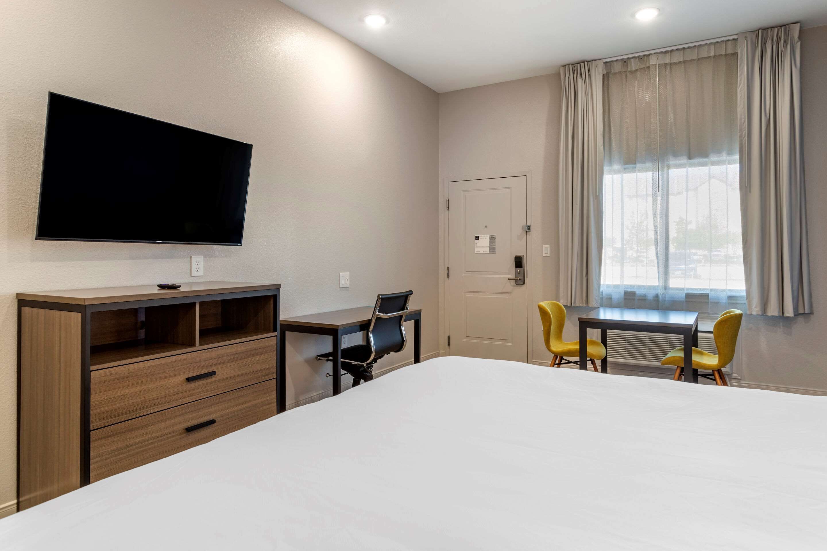 A hotel room with a bed, a desk with a chair, a television mounted on the wall, and a window with curtains.