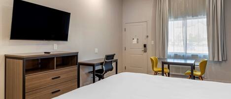 A hotel room with a bed, a desk with a chair, a television mounted on the wall, and a window with curtains.
