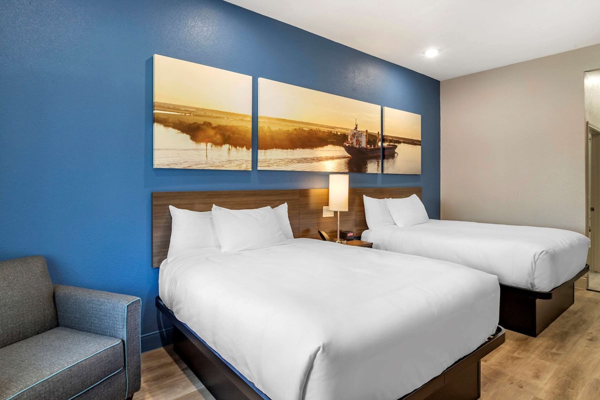 Ein Hotelzimmer mit zwei Betten, einem Schlafsofa, einem Wandbild und einem Nachttisch mit Lampe.