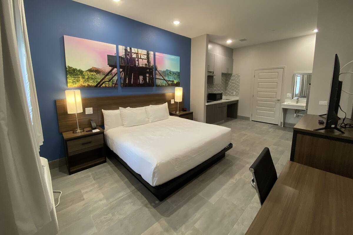 Ein modernes Hotelzimmer mit einem großen Bett, Nachttischen inklusive Lampen, einem Schreibtisch mit Computer, einer Küchenzeile und einem Badezimmer.