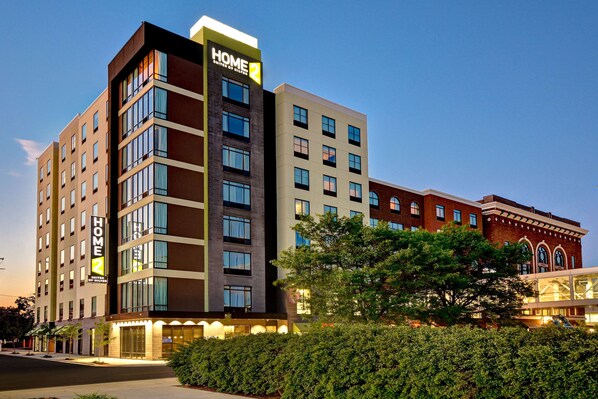 Un hotel de varios pisos con un llamativo letrero de 'Home 2', amplias ventanas y un edificio clásico en el fondo.