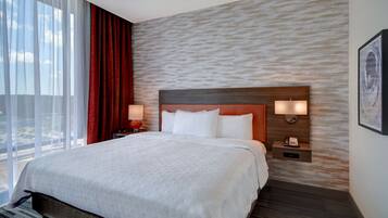 Habitación de hotel con una cama grande, cabecera con iluminación integrada, mesita de noche con lámpara y vistas al exterior a través de amplios ventanales.