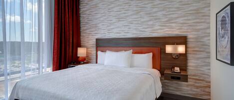 Habitación de hotel con una cama grande, cabecera con iluminación integrada, mesita de noche con lámpara y vistas al exterior a través de amplios ventanales.