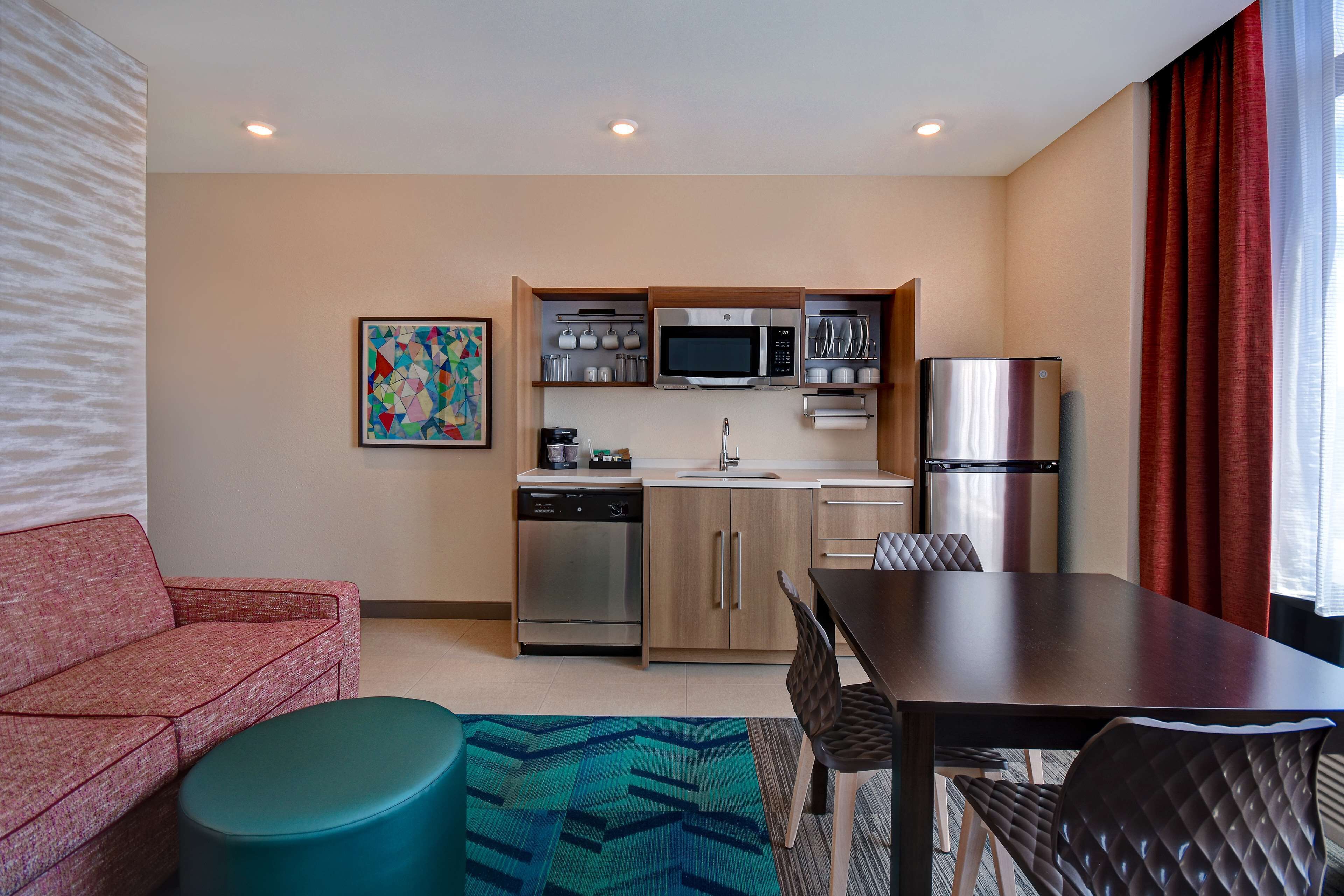 A compact hotel room with a kitchenette, a sofa, a dining table, and a patterned rug.