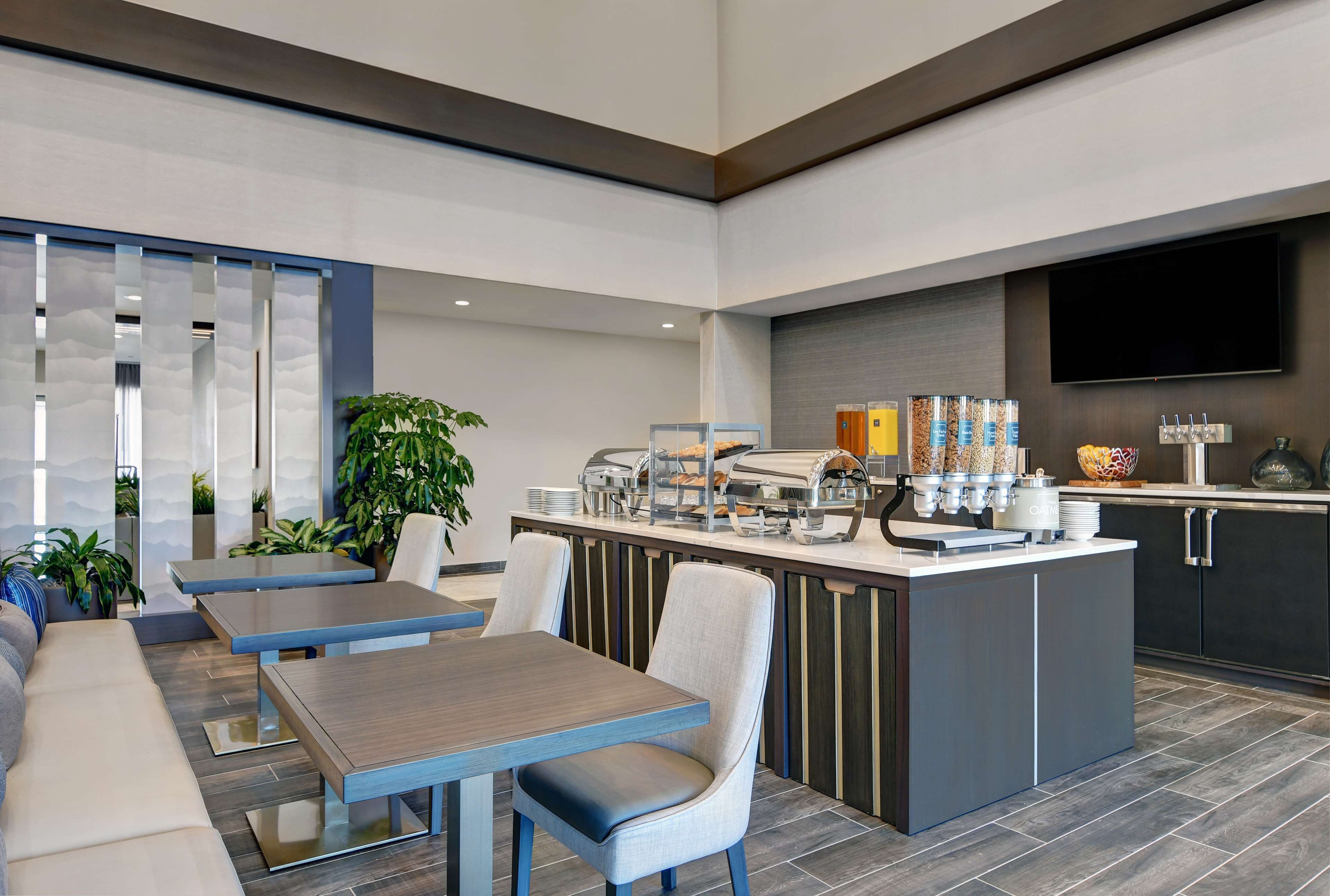 Un moderno vestíbulo de hotel con zona de comedor, una encimera de cocina con alimentos para el desayuno y un televisor de pantalla plana en la pared.