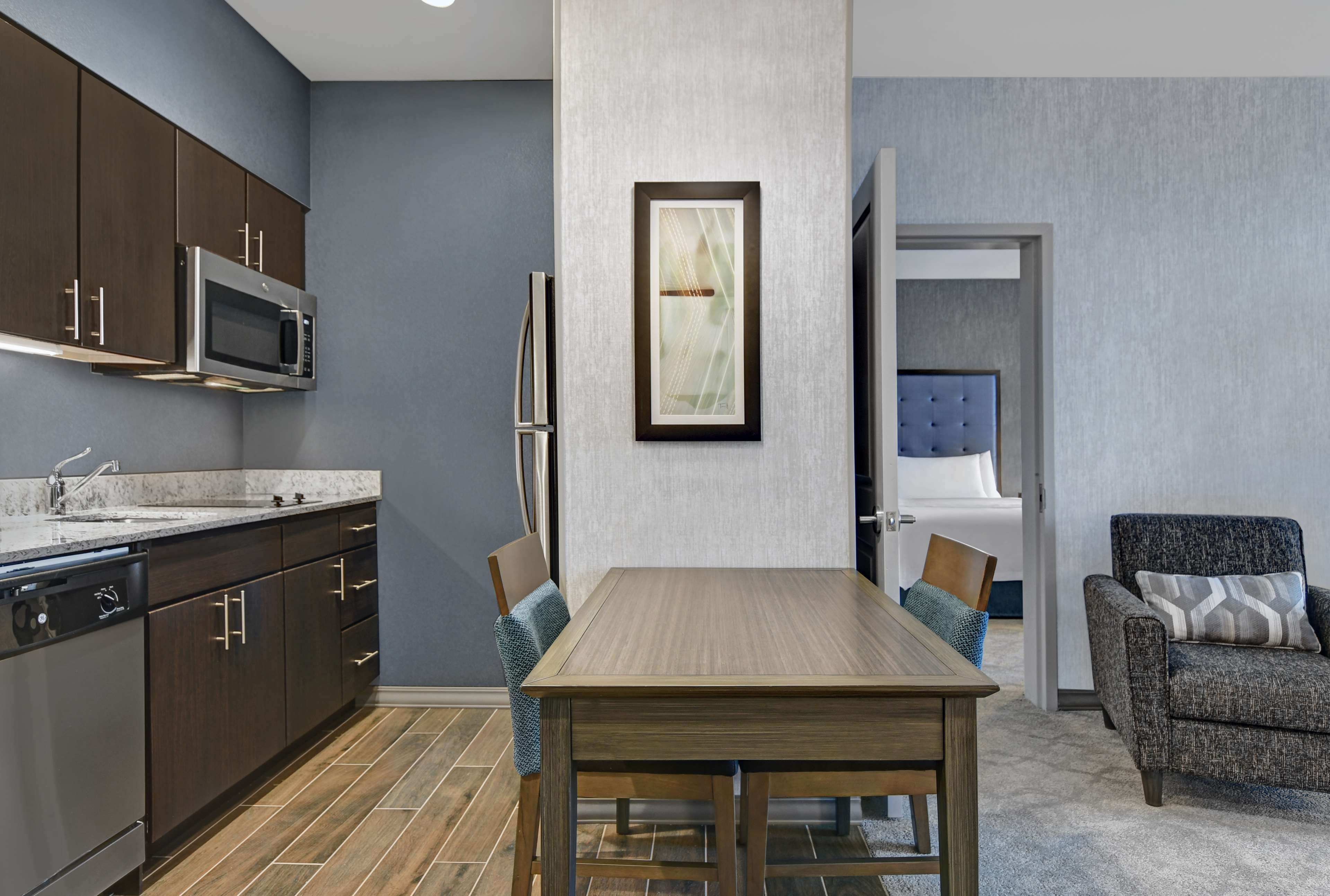 Uma cozinha moderna com armários escuros, micro-ondas e lava-louças. Há uma mesa de jantar com cadeiras e um quadro na parede.