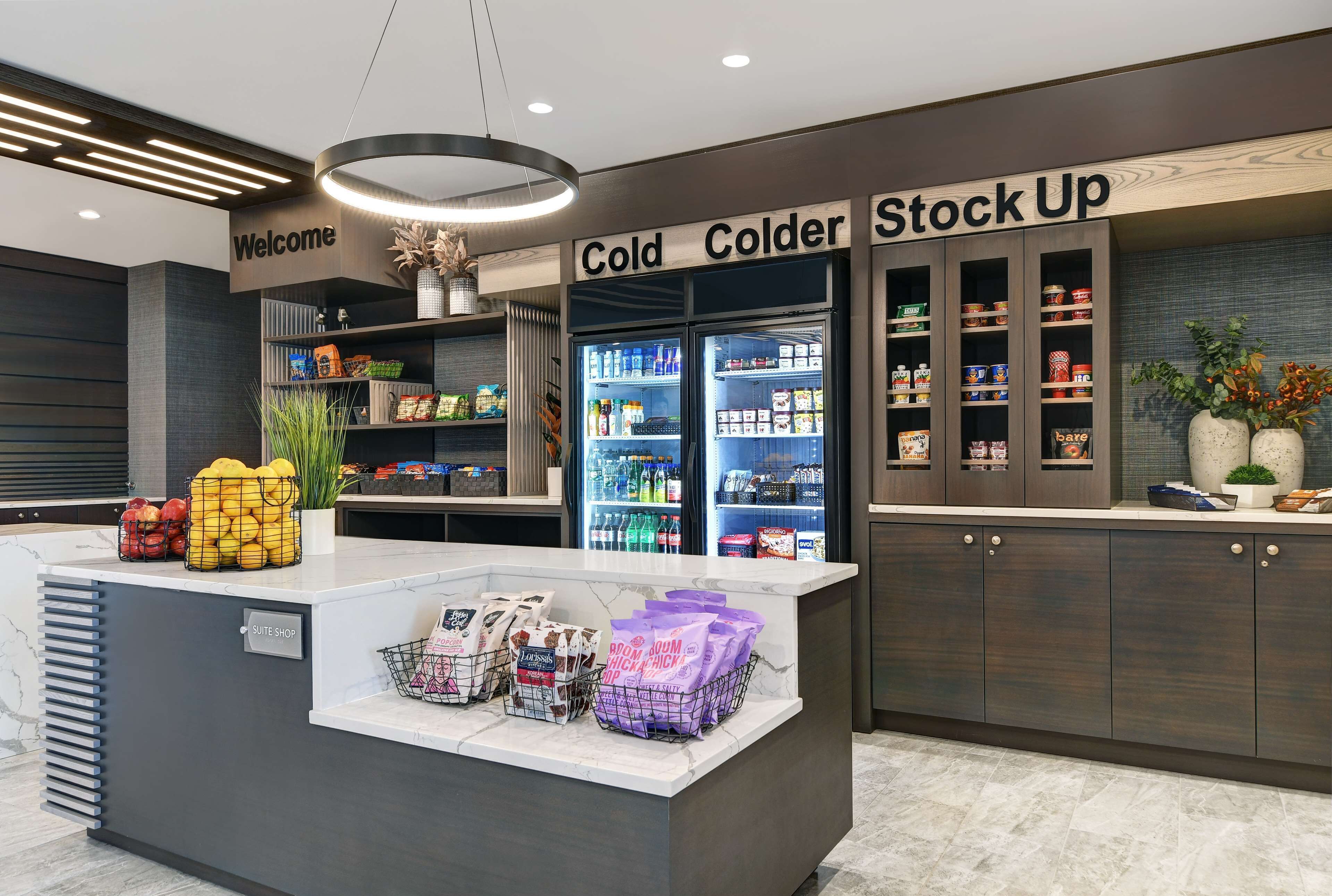 A modern hotel lobby with a reception desk, a 'Welcome' sign, and a 'Cold, Cooler, Stock Up' section with refrigerated display cases and shelves.