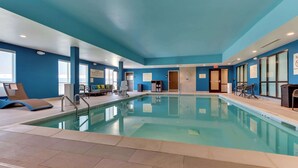 Une piscine intérieure à l’eau claire, entourée de murs bleus et surmontée d’un plafond. On y trouve des chaises longues et un petit coin salon avec une table et des chaises.