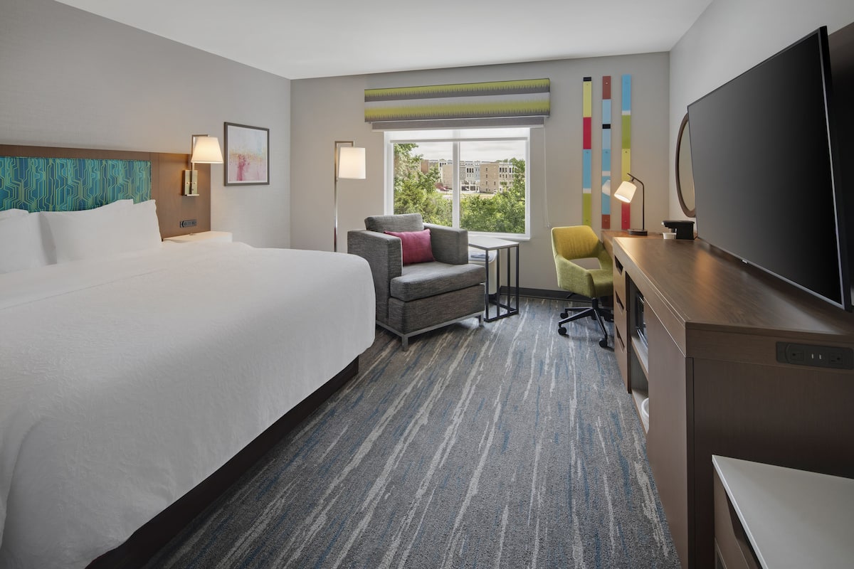 Ein Hotelzimmer mit einem großen Bett, einem Schreibtisch mit Fernseher, einem Sessel und einem Fenster mit Blick auf Gebäude.