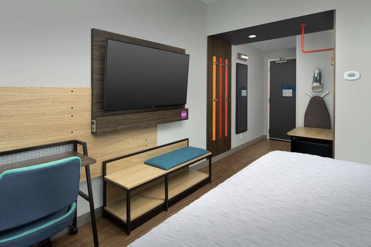 A modern hotel room with a large bed, a desk with a chair, a TV mounted on the wall, and a bathroom visible through an open door.