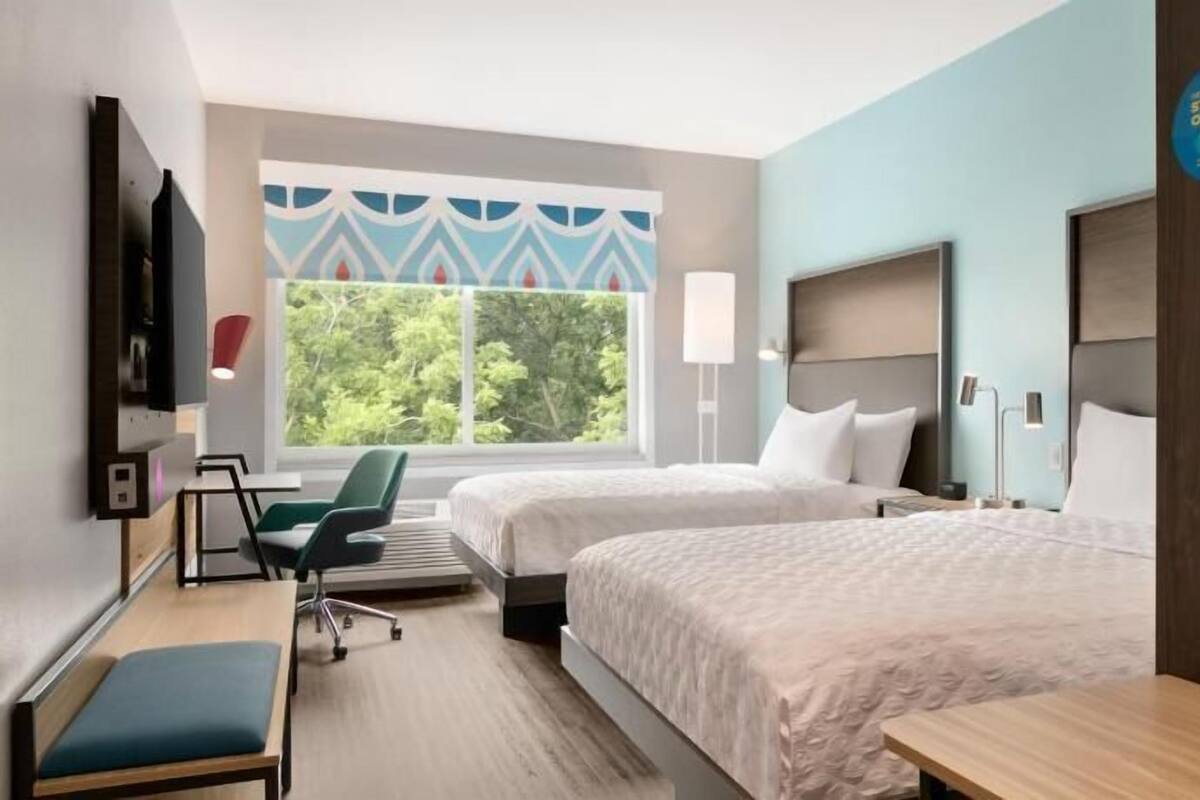 A hotel room with two beds, a desk, a chair, and a large window offering a view of greenery.