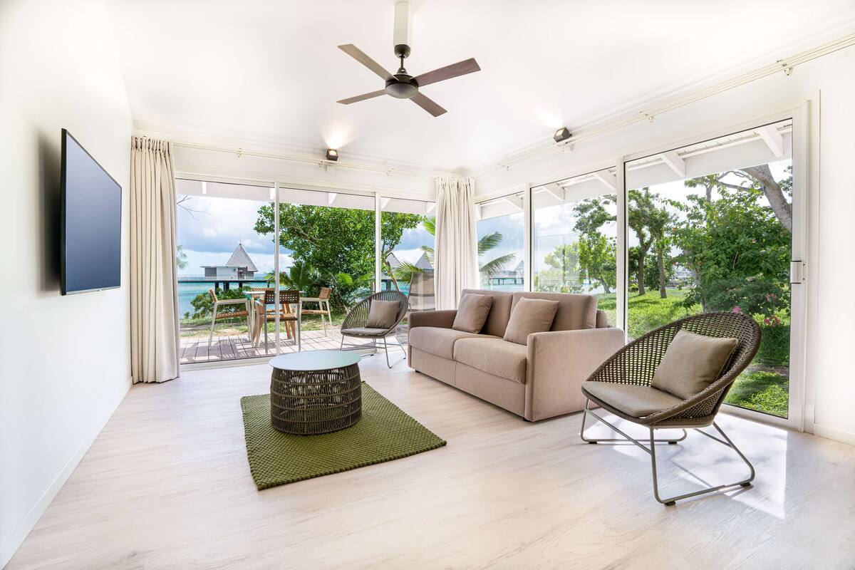 A spacious living room with a sofa, armchair, and coffee table, large windows with a view of the sea, and a ceiling fan.