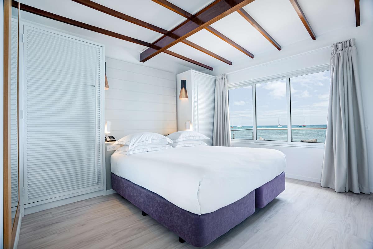 A modern bedroom with a large bed, a view of the sea, and wooden beams on the ceiling.