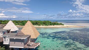 Een overwaterbungalow met rieten dak, helder turquoise water en een strand op de achtergrond.