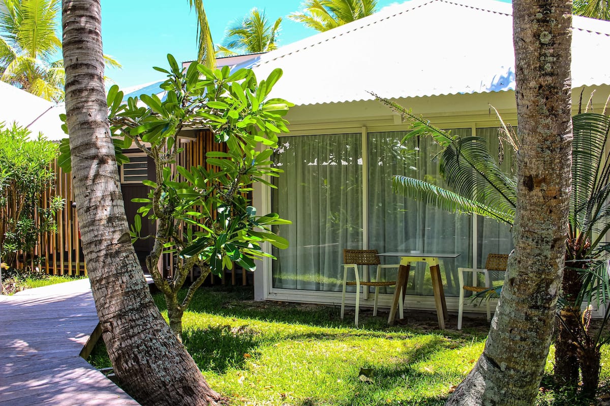 A covered patio with a table and chairs, surrounded by palm trees and a wooden fence.