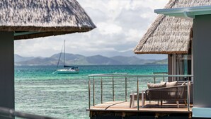 Bungalows sobre el agua con techos de paja, asientos en terraza de madera y vistas al océano y un velero.