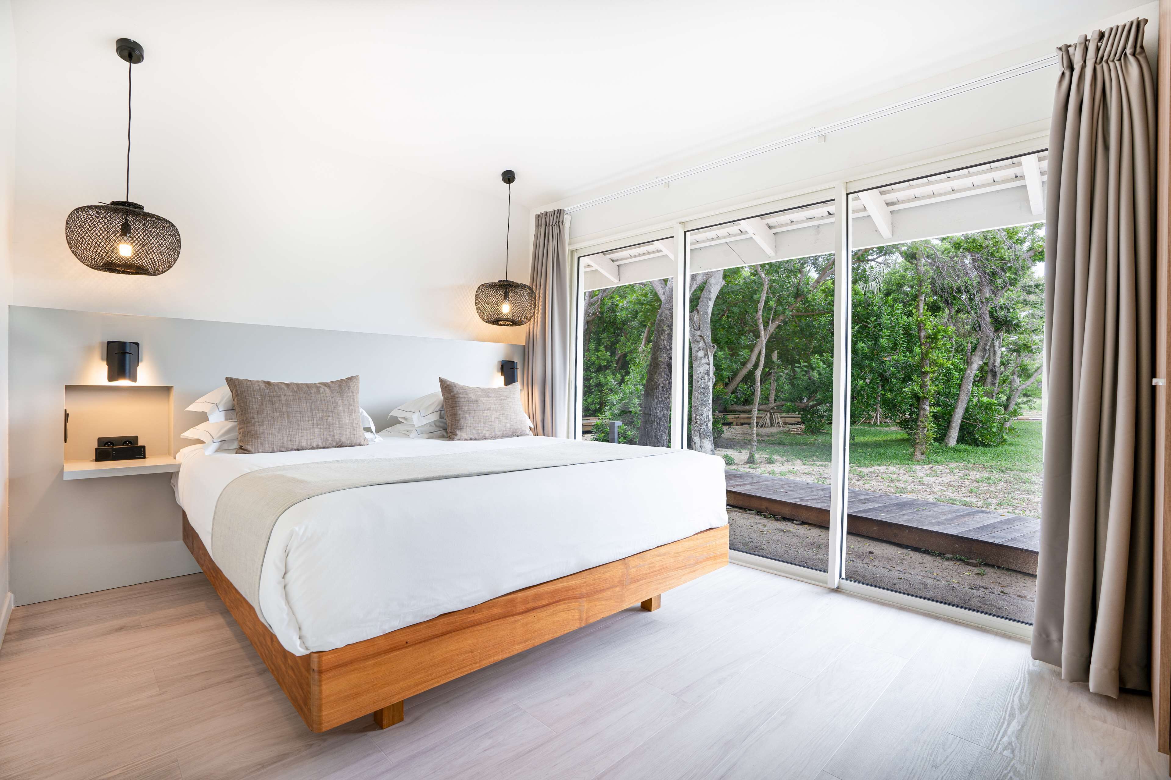 A modern bedroom with a large bed, a nightstand, and a window with a view of greenery.