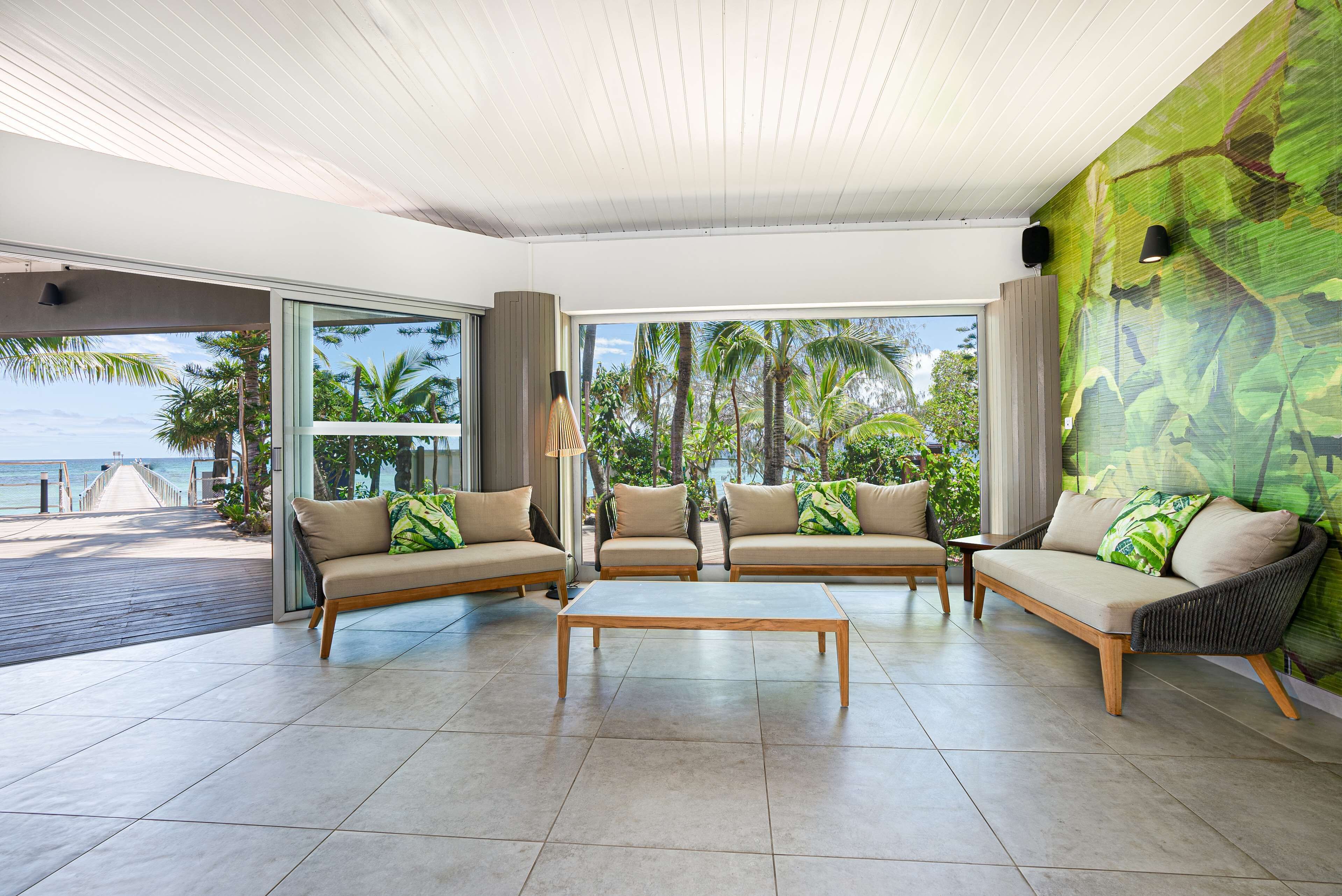 A spacious living area with a large window overlooking a tropical beach. There are three sofas and a coffee table.