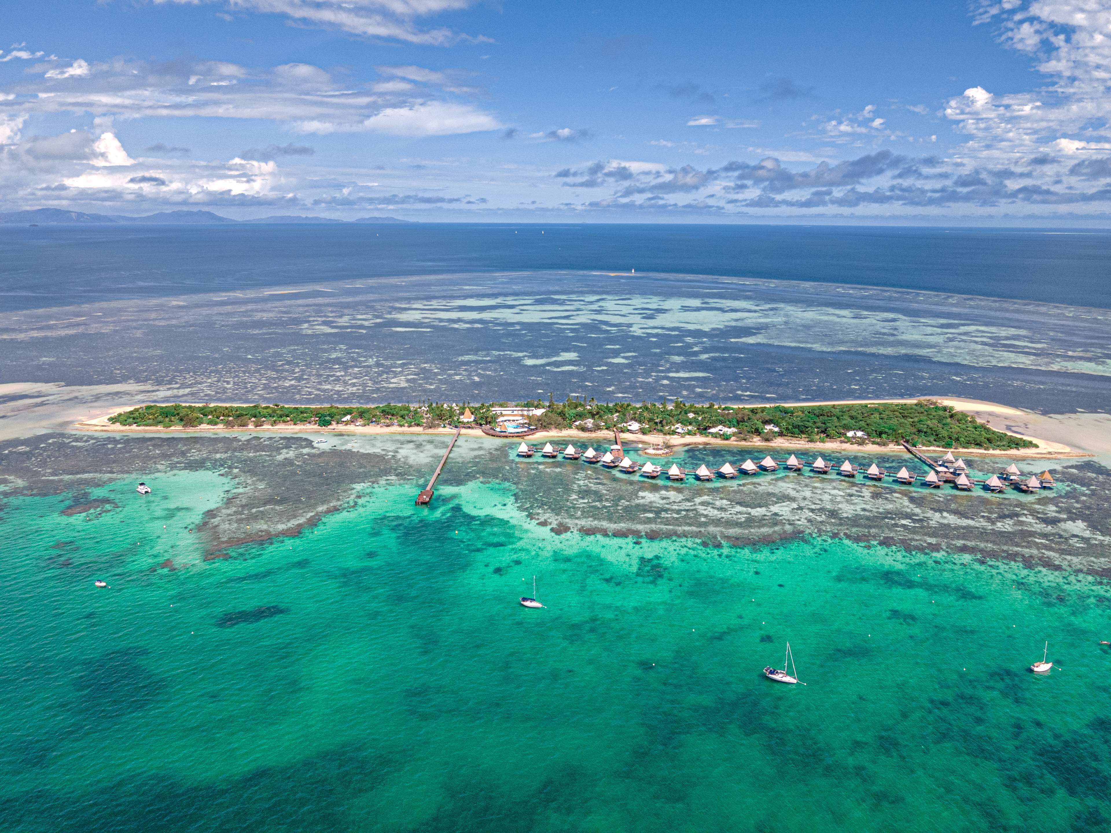 Vista aérea de un resort tropical con bungalós sobre el agua, aguas turquesas cristalinas y una cadena montañosa distante.