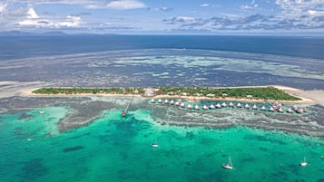 Vista aérea de un resort tropical con bungalós sobre el agua, aguas turquesas cristalinas y una cadena montañosa distante.