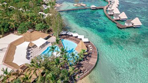 Vista aérea de un resort con bungalows sobre el agua unidos por pasarelas, un área de piscina y abundante vegetación.