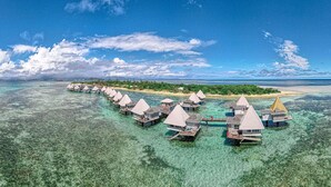 Vista aérea de bungalós sobre agua con aguas azul cristalina y una isla distante.