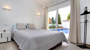 A bedroom with a bed, a bedside table, a lamp, and a view of a swimming pool.