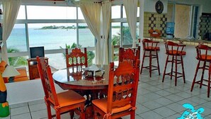 Zona pranzo con vista sul mare, sedie di legno, un tavolo rotondo e angolo cottura con rivestimento in piastrelle.