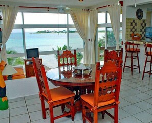 A dining area with a view of the sea, wooden chairs, a round table, and a kitchen area with a tiled backsplash.