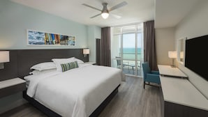 Quarto de hotel com uma cama grande, uma escrivaninha, uma cadeira, uma TV e uma varanda com vista para o oceano.