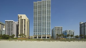 Hotel alto com vários andares, uma praia em primeiro plano e palmeiras.