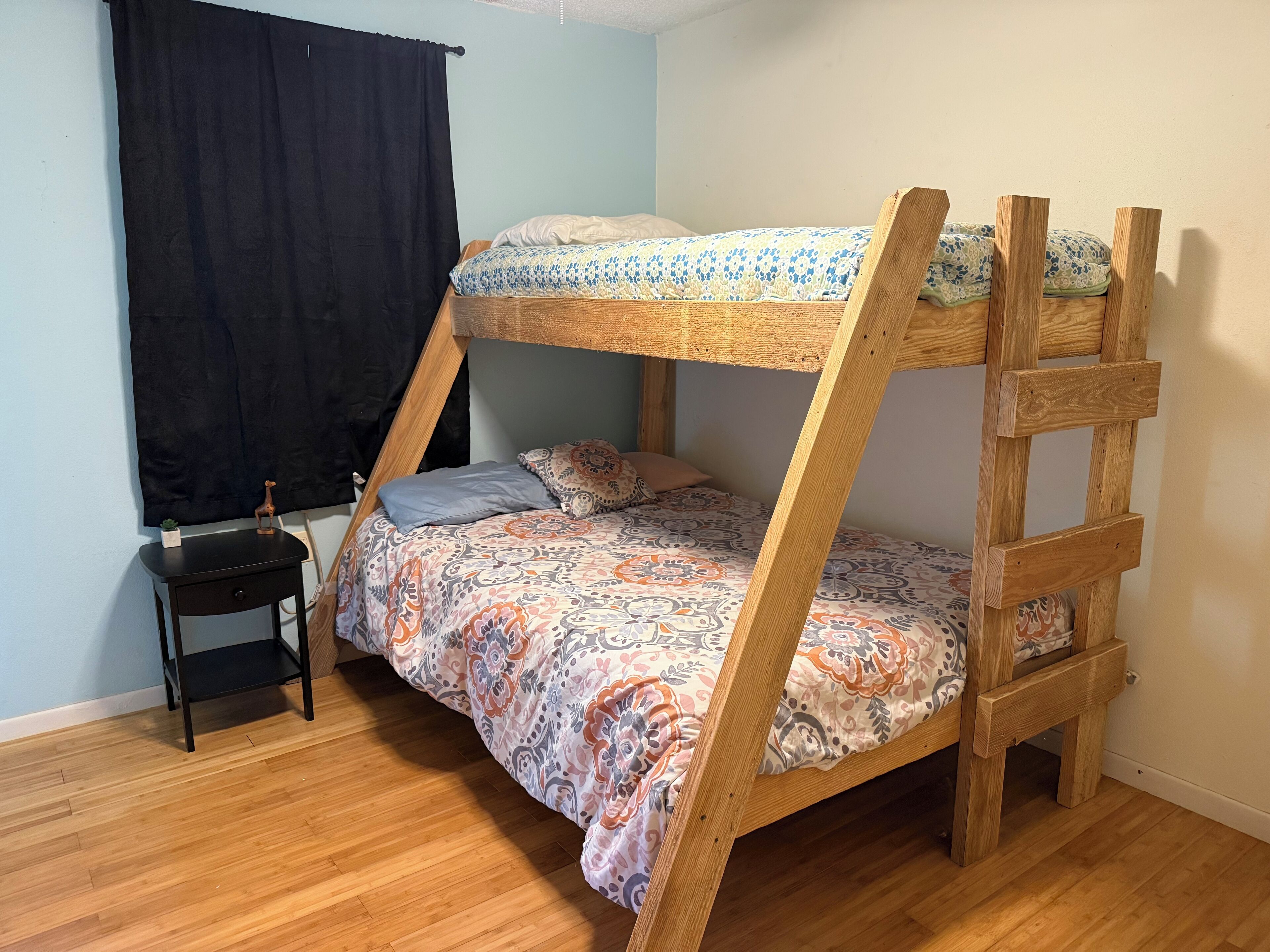 A bunk bed with a patterned bedspread, a small bedside table with a lamp, and a black curtain.