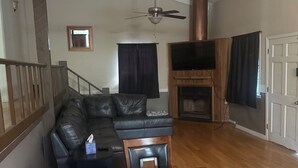 Un salon comprenant un canapé d’angle en cuir noir, une table à manger en bois avec des chaises, un ventilateur de plafond, une cheminée et un téléviseur fixé au mur.